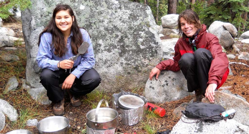 Two people kneel beside cooking pots sitting on the ground and smile for the photo